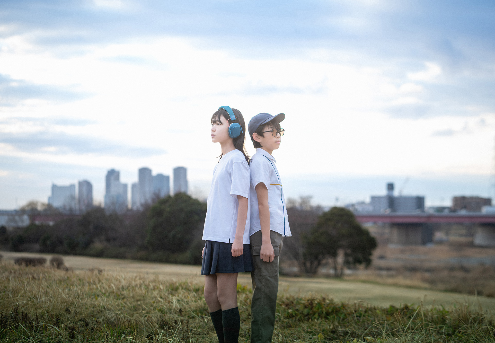 河川敷で背中合わせに立つ、ヘッドフォンをした少女と少年